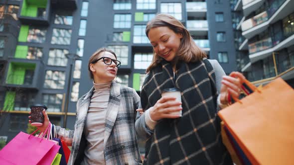 Two Happy Women Walk with Shopping Bags and Takeaway Coffee After a Successful Shopping and Talk alt