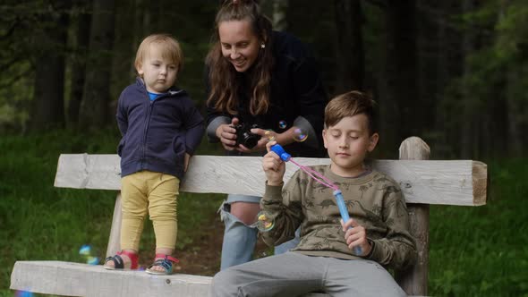Young mother with her sons playing with soap bubbles and makes pictures on the camera in the park. alt