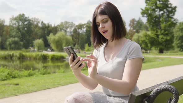 A Young Caucasian Woman Works on a Smartphone and Acts Upset As She Sits on a Bench alt