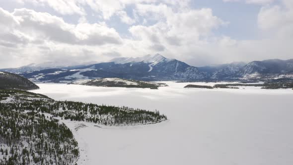 Aerial moving backward to reveal icy, frozen lake surrounded by snow and green pine trees with large alt