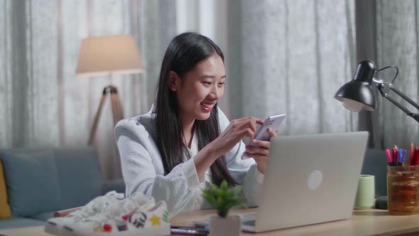 Female Footwear Designer Looking At The Shoes And Using Smartphone While Working On A Laptop At Home alt