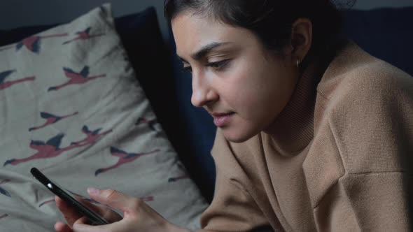 Side View Close Up Of A Young Indian Woman Lying On A Bed Using A Smartphone Reading The News alt