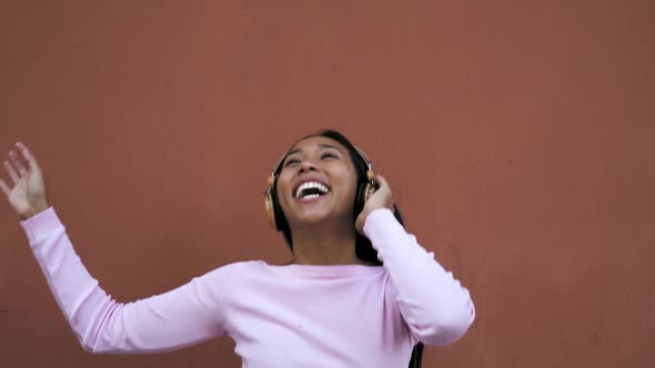 Happy young Latin woman having fun dancing and listening music with wireless headphones alt