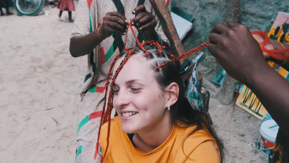 African Woman Weaving African Braids with Red Kanekalon Outdoor Zanzibar Africa alt