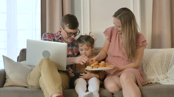 Father Works From Home Remotely During Quarantine on a Laptop with Child on the Couch, Wife Brings alt