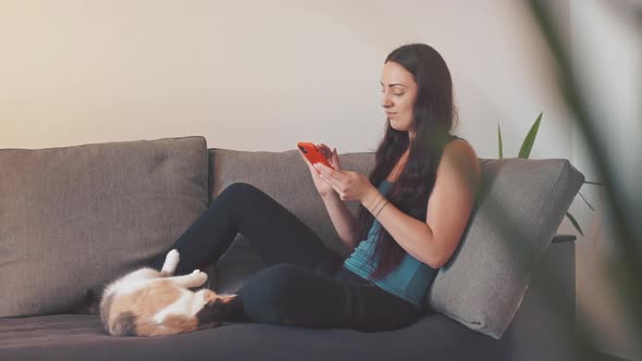 Young Woman Sit on Sofa with Cat at Home and Using Smartphone