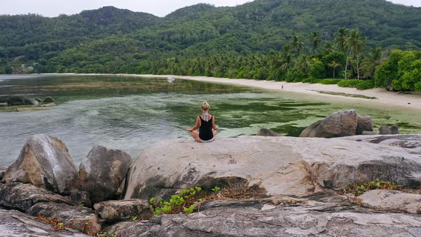 Aerial Circle Footage Around Blonde Tanned Girl Sitting in Yoga Pose and Meditating on Granite Rock alt