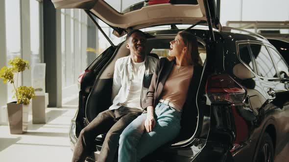 Positive Multiracial Couple Sitting in Hugs in Open Trunk of Their New Auto alt