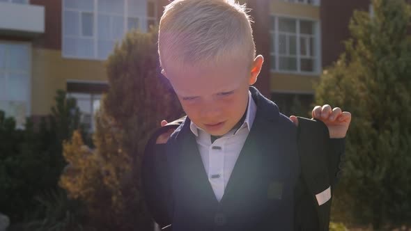 A Little Boy in a Shirt and Jacket with a School Bag Near the School Building alt