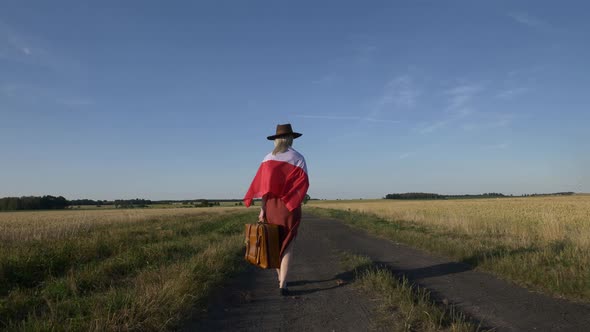 Girl in Poland flag with suitcase on country road in sunset