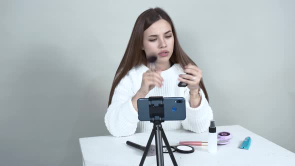 A Caucasian Girl Blogger in Front of a Cellphone Puts a Blush on Her Face Brush. alt