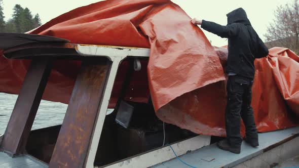 Young man untying  waterproof cover of wooden boat before starting restoration maintenance alt