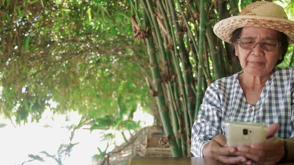 Asian elderly woman wearing a hat and eyeglasses looking at a smartphone Online video call alt
