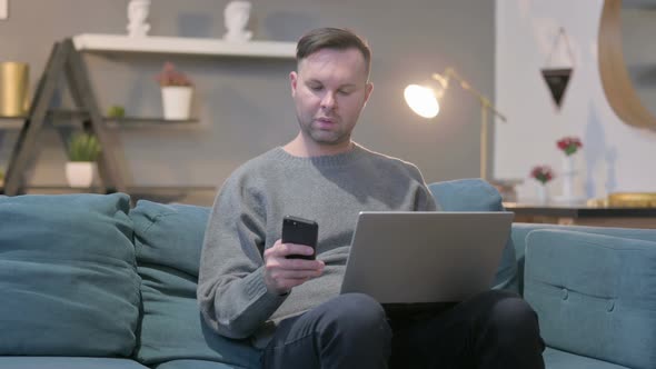 Casual Man with Laptop Using Smartphone on Sofa alt