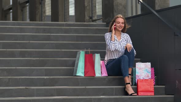 Girl Sitting on Stairs with Bags Talking on Mobile Phone About Sale in Shopping Mall in Black Friday alt