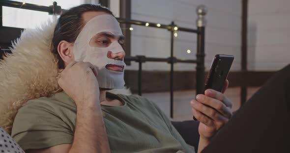 A Man in a Tissue Moisturizing Mask is Resting on a Bed or Sofa with a Phone
