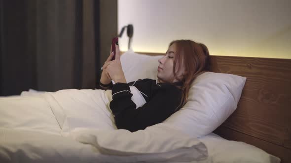 Side View of Relaxed Beautiful Redhead Girl Lying in Bed and Using Social Media on Smartphone alt