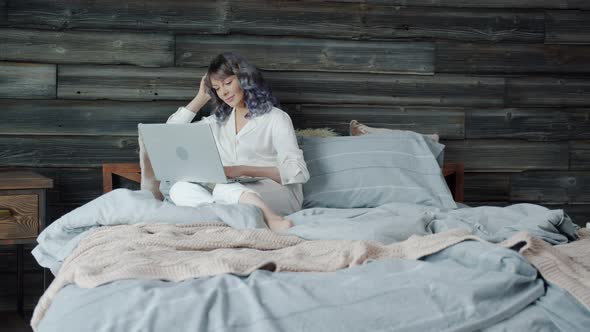 Slow Motion of Cheerful Young Woman Using Laptop in Bed and Smiling Enjoying Internet Content alt