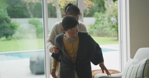 Senior asian mother being helped to armchair by adult daughter in living room alt