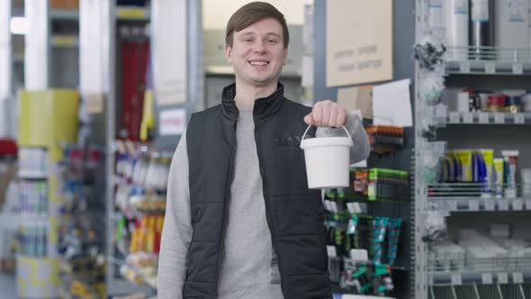 Positive Male Seller with Toothy Smile Stretching Paint Can to Camera Standing in Hardware Store alt