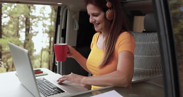 Senior caucasian woman using laptop and wireless headphone inside mini van camper alt