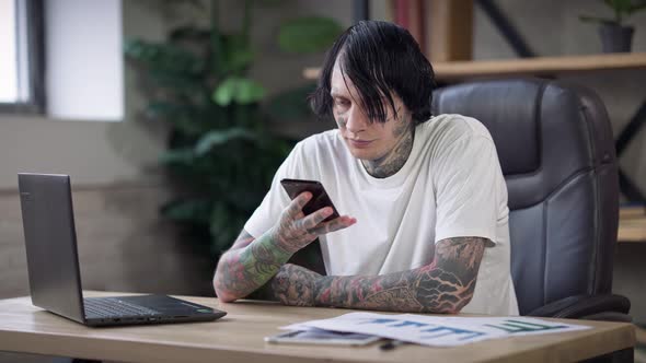 Focused Male Tattooed Freelancer Sitting at Table with Graphs and Laptop Surfing Internet on alt