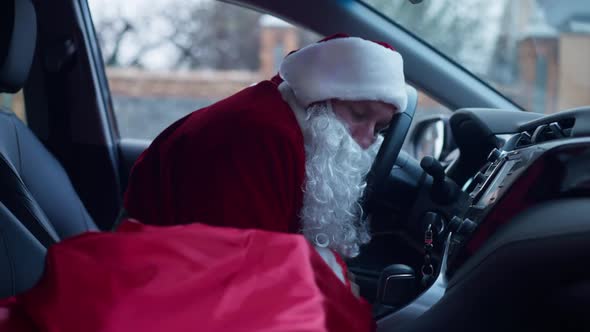 Side View Exhausted Father Christmas Sleeping in Car Yawning Leaning on Steering Wheel alt