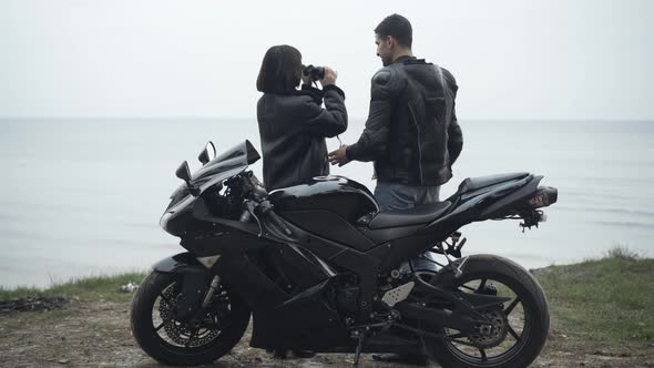 Back View of Couple of Young Bikers Standing on River Bank and Looking Away Through Binoculars. Wide alt