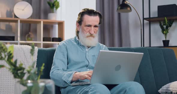 Senior Bearded Man Sitting on the Soft Couch in Cozy Apartment and Typing on Computer alt