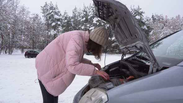A Girl in a Pink Winter Jacket and a White Hat Inspects a Broken Car Under the Hood in Winter alt