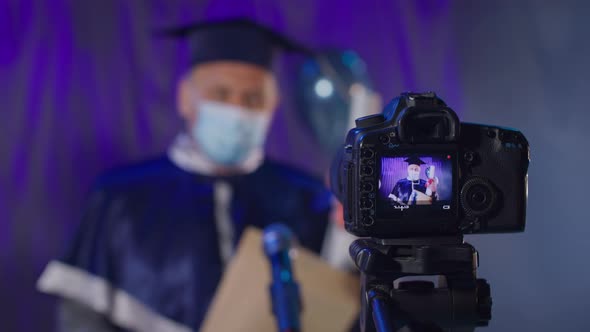 Male Professor in Gown and Medical Mask Solemnly Greets End of School Year and Presents Diplomas Via
