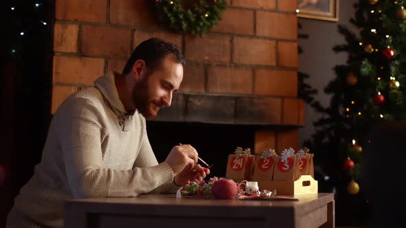 Medium Shot of Focused Young Man Writing Number on Red Bag By Brush and White Paint Sitting at Table alt