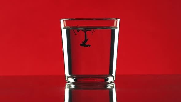 Drop of Black Ink Dripping Into Clear Water in Glass Standing on Red Background in Slowmotion alt