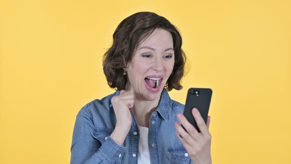 Old Woman Celebrating on Smartphone, Yellow Background  alt