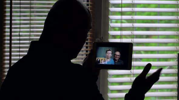 A Bald Man Stands Against the Light at a Window with Blinds, She Is Talking on a Video Conference on alt