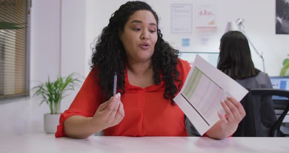 Happy biracial businesswoman sitting at desk, holding documents, making video call in modern office alt