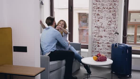 Happy Cheerful Modern Couple which Sitting on Sofa in Hotel Room and Hugging Each Ather alt