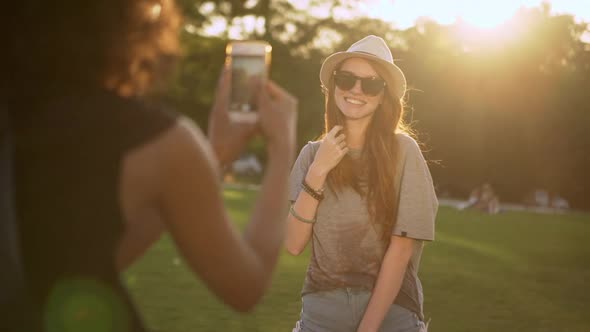 Pretty Ginger Young Girl in Hat and Sunglasses Posing While Her Tanned Friend Taking Photo of Her in alt