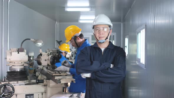 Factory worker with white hardhat and blue uniform stand with arm-crossed in front of co-worker alt