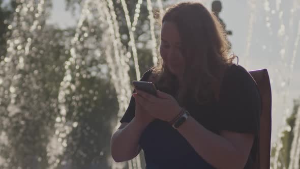 Portrait of Pretty Woman with a Phone in Her Hands Standing Near the Fountain