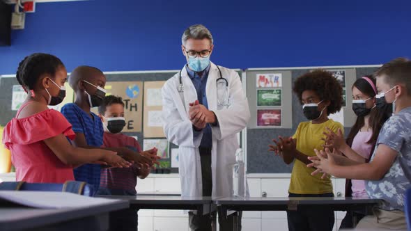 Diverse schoolteacher and schoolchildren standing disinfecting hands, all wearing face masks alt