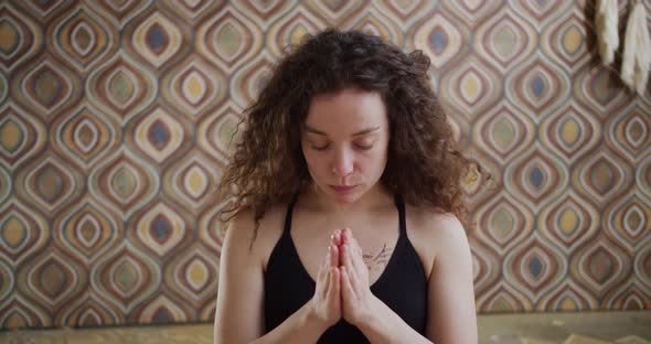 Relaxed CurlyHaired Young Woman in Sportswear is Meditating in Position on Yoga Mat with Closed Eyes