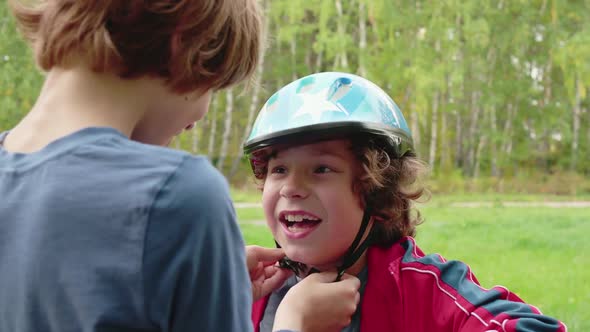 Older Brother Putting Bicycle Helmet on Little Brother Head in Summer Park alt