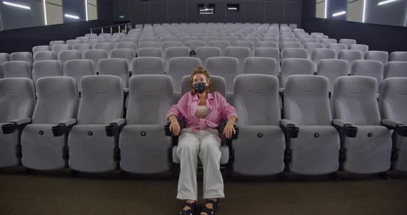 a Young Stylish Woman in a Protective Face Mask is Sitting on the First Row of an Empty Cinema Hall alt