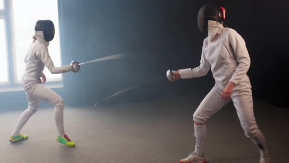 Two Young Women Having an Intense Training in a Fencing Duel in the Dark Smoky Studio alt