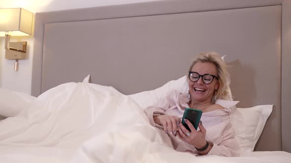 A Blonde Woman with Glasses is in the Bedroom Lying on the Bed with White Rumpled Bed Linen alt
