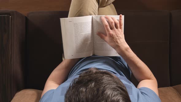 Young adult man reads a book on a sofa at home