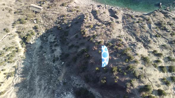Aerial Drone View of a Man Flying a White and Blue Paraglider Over a Hill and Trees to the Sea Waves alt