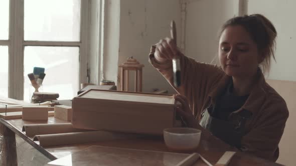 Young Woman Applying Protective Varnish on Wood Handicraft alt