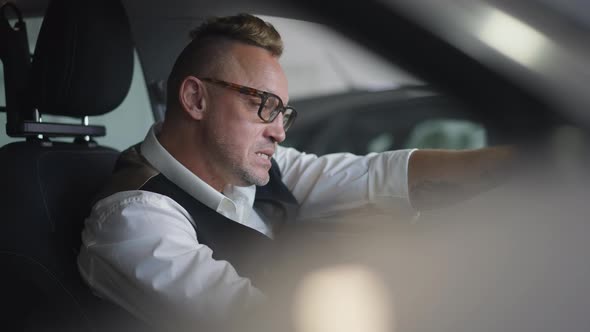 Tattooed Adult Caucasian Man in Eyeglasses with Mohawk Haircut Sitting on Driver's Seat in Car alt